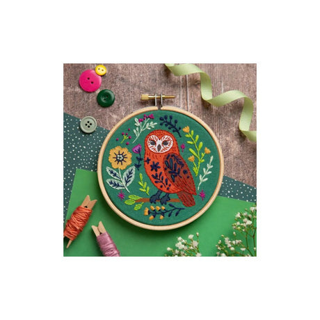 A circular embroidery hoop containing a colorful owl and floral design stitched on green fabric, displayed on a wood surface with buttons, ribbon, spools of thread, and small flowers around it.