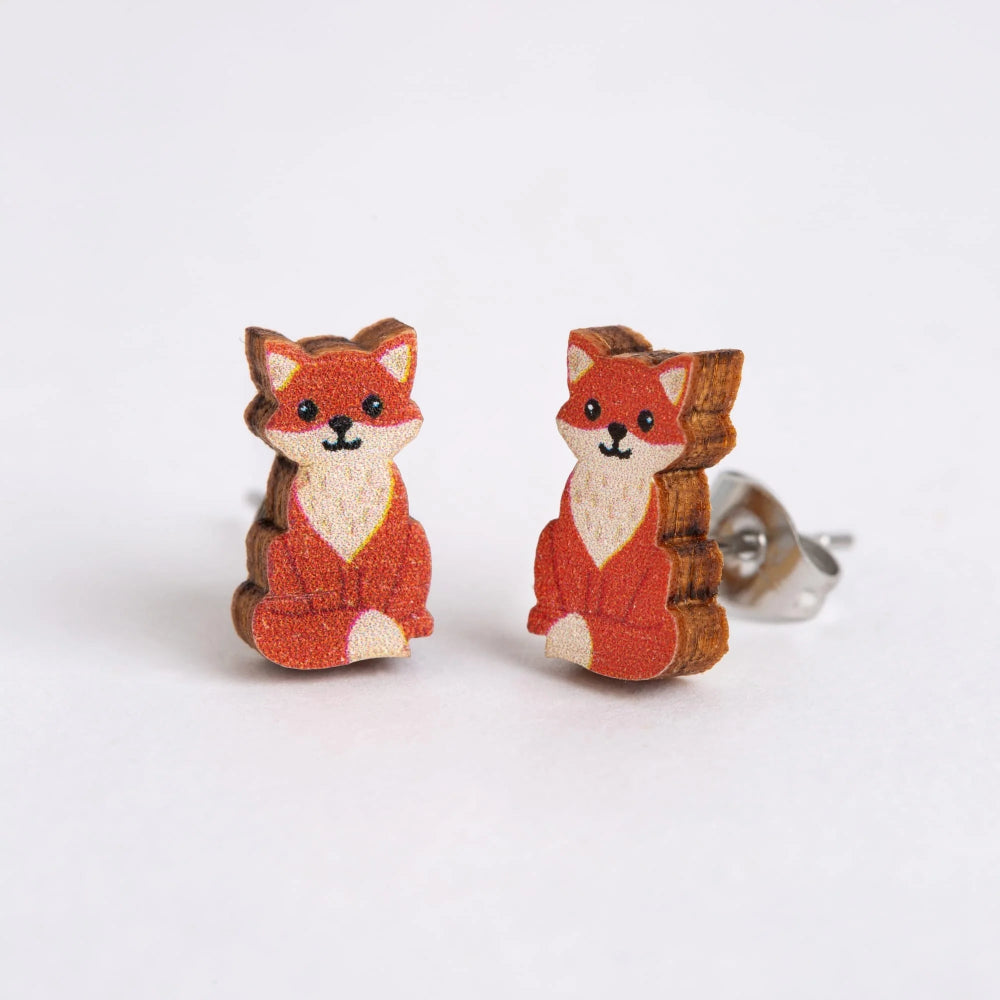 A pair of wooden stud earrings shaped like cute sitting foxes, featuring orange-brown fur, cream-colored faces and chests, and silver-tone post backings, set against a plain white background.