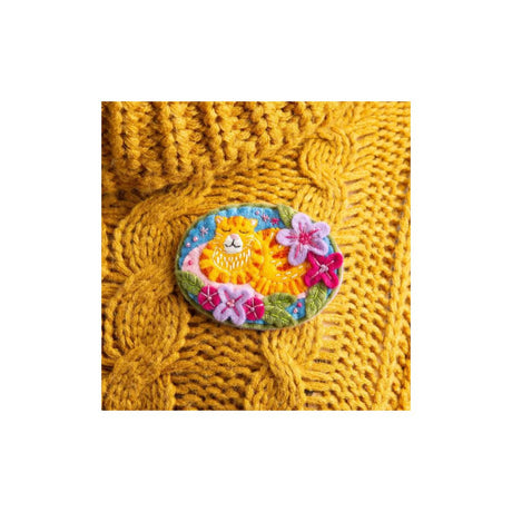 An oval embroidered patch featuring a yellow cat surrounded by pink and purple flowers on a blue background, displayed on a yellow cable-knit sweater against a white backdrop.