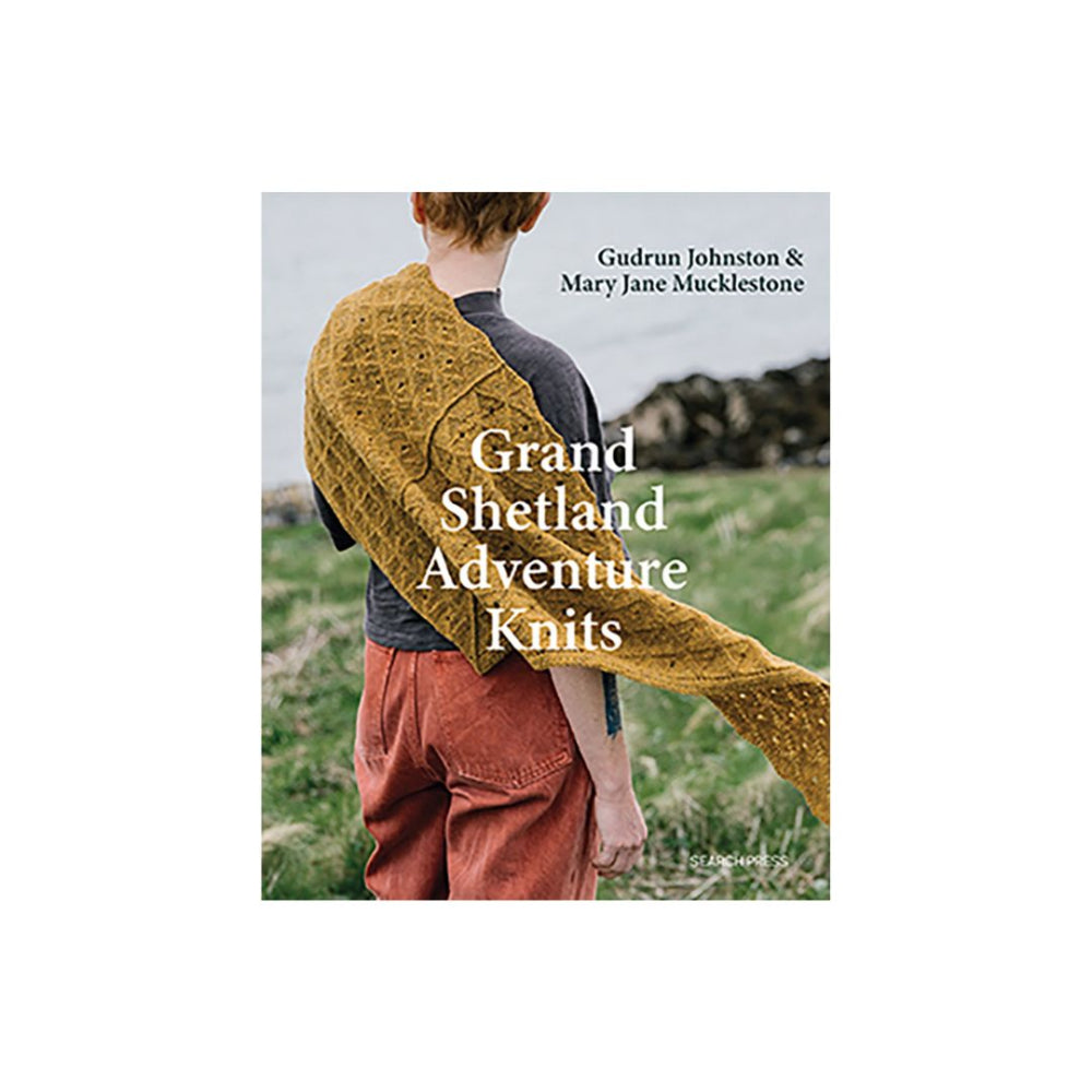 Book cover for 'Grand Shetland Adventure Knits' by Gudrun Johnston & Mary Jane Mucklestone, showing a person outdoors wearing and draping a mustard yellow knitted shawl over grassy ground.