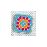 A square fabric patch with rounded corners featuring a colorful geometric granny square pattern, displayed on a white textured mesh background.