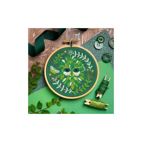 A circular embroidery hoop containing a green tree-of-life design with leaves and a butterfly, surrounded by green thread spools, buttons, ribbon, and small green leaves on a styled flat-lay background.