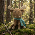 A knitted moose toy with brown and beige knit body, cream-colored antlers and hooves, and a teal knit scarf