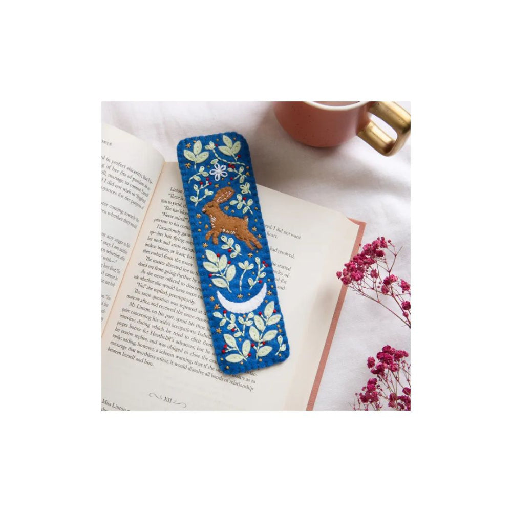 A blue fabric bookmark with a embroidered hare, moon, and floral pattern, resting on open book pages with a pink mug and dried flowers in the background.