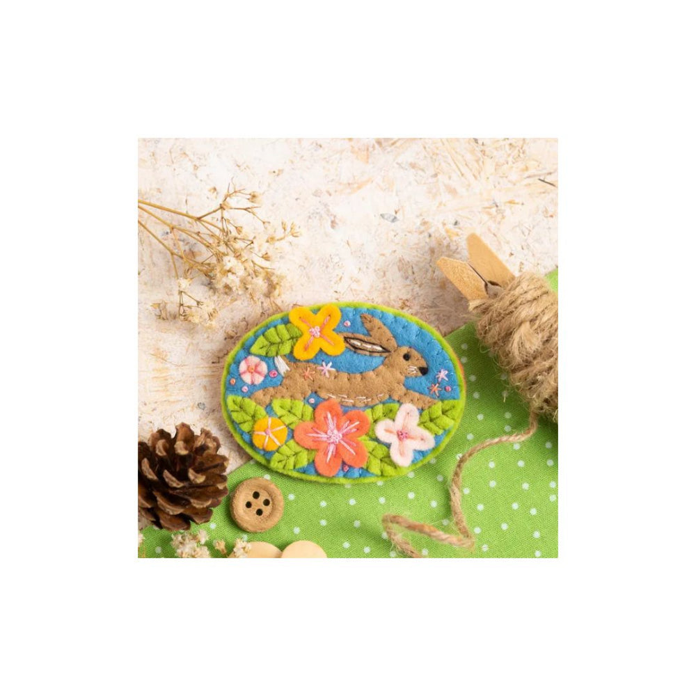 An oval felt patch featuring a brown bunny among colourful flowers on a green background, styled on a light surface with a pinecone, button, twine, and dried flowers.