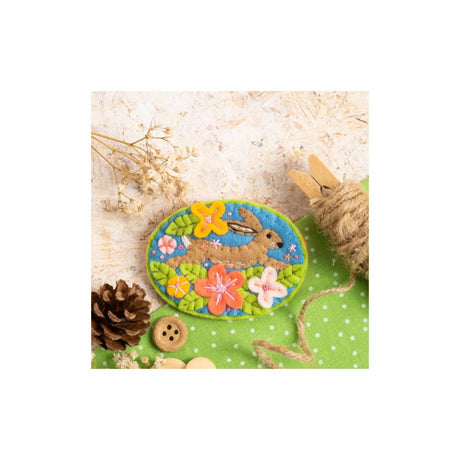 An oval felt patch featuring a brown bunny among colourful flowers on a green background, styled on a light surface with a pinecone, button, twine, and dried flowers.