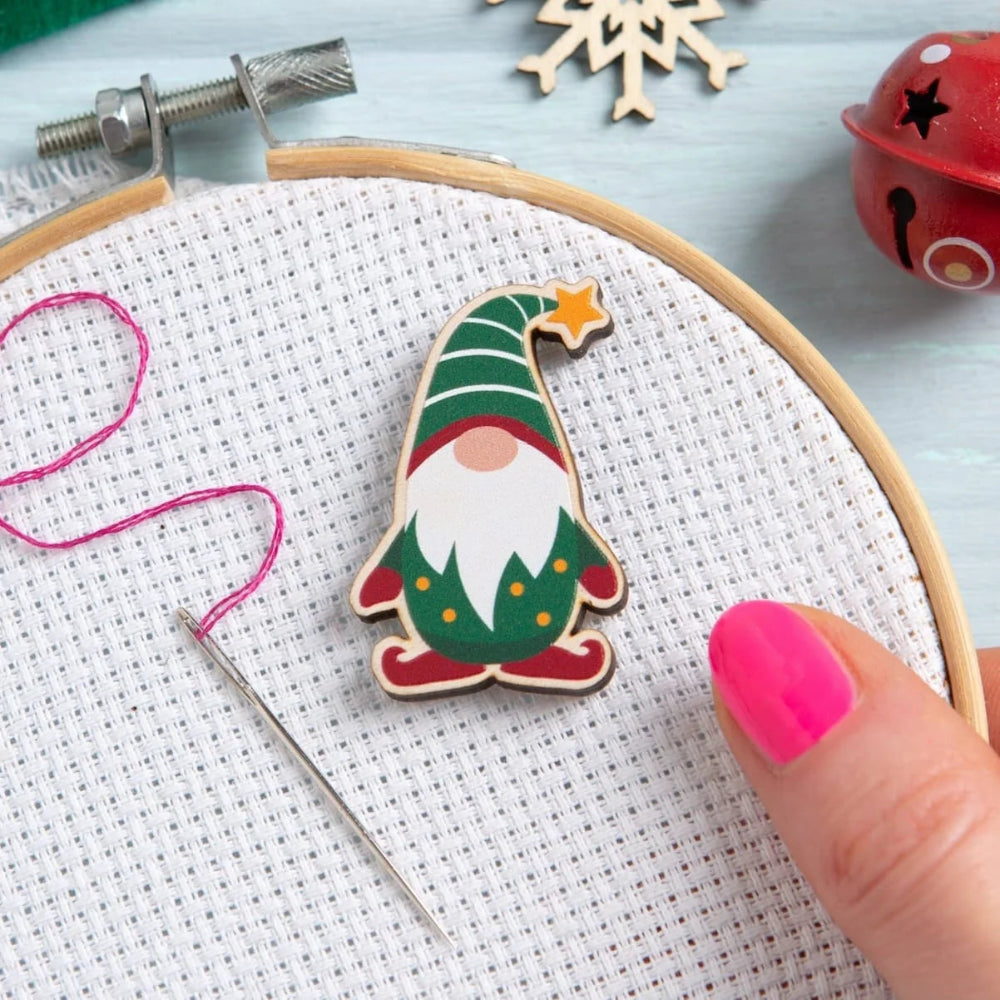 A Christmas gnome needle minder with a green striped hat, red shoes, white beard, and yellow star, shown inside a white embroidery hoop with a needle and pink thread. A hand with bright pink nail polish is visible on the right.