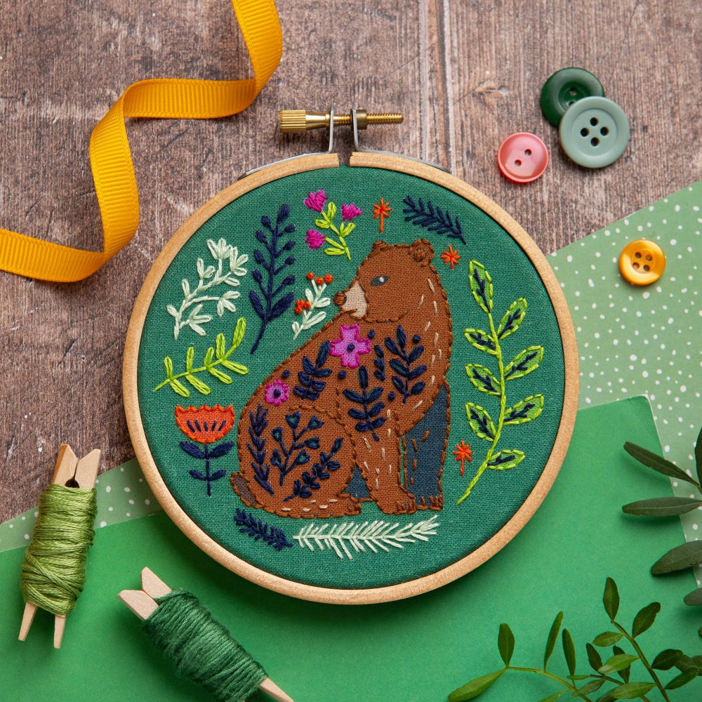 Embroidery hoop with a bear and floral design on a wooden surface with buttons and thread.