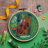 Embroidery hoop with a bear and floral design on a wooden surface with buttons and thread.
