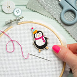 A close-up of a hand with pink nail polish holding an embroidery hoop with white Aida fabric, a wooden needle, pink thread, and a small embroidered penguin patch wearing a party hat and pink scarf. Scissors, buttons, and textured paper are arranged on a pale mint background.
