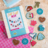 A felt craft kit box labelled Hawthorn Handmade Winter Garland is shown on a pink polka-dot sheet with several completed felt patches and garments arranged around it on a light wood surface.