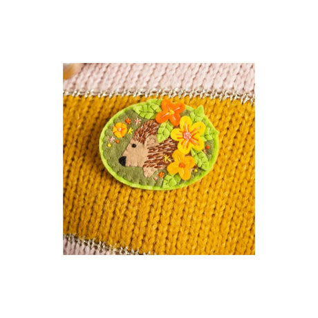 An oval embroidered patch featuring a brown hedgehog surrounded by orange and yellow flowers on a green background, sewn onto a mustard yellow knitted garment.