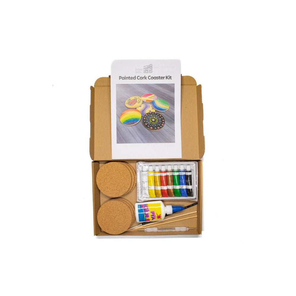 A painted cork coaster kit displayed in an open brown cardboard box on a white background. The kit includes two round cork coasters, a small bottle of acrylic paint, wooden sticks, a paintbrush, and a printed instruction sheet showing painted examples.