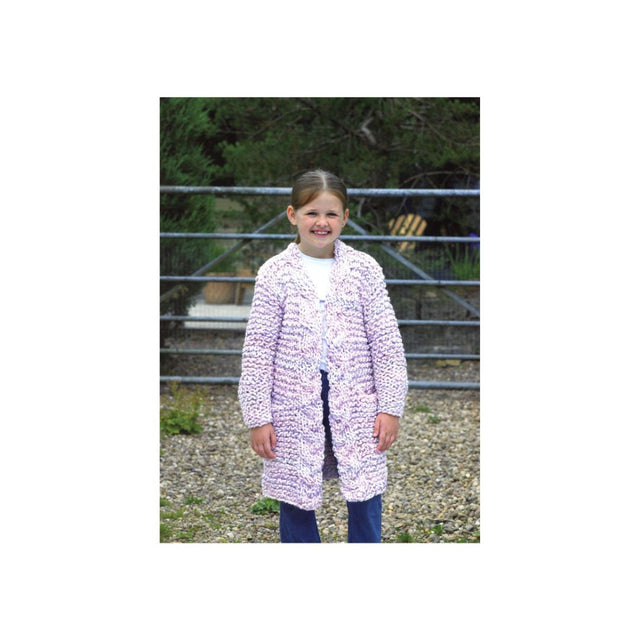 A young girl stands outdoors in front of a metal gate and gravel ground, wearing a long, chunky knit cardigan with a pink and white abstract pattern over a white top and dark blue jeans.