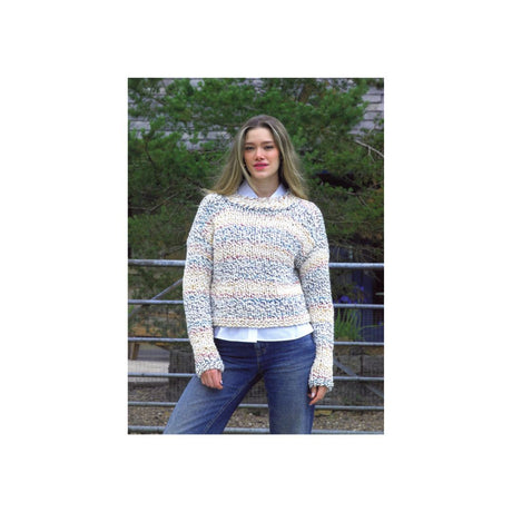 A woman wearing a chunky knit sweater with horizontal stripes of cream, light blue, and beige, layered over a white collared shirt and blue jeans, standing outdoors in front of a metal fence and green trees.