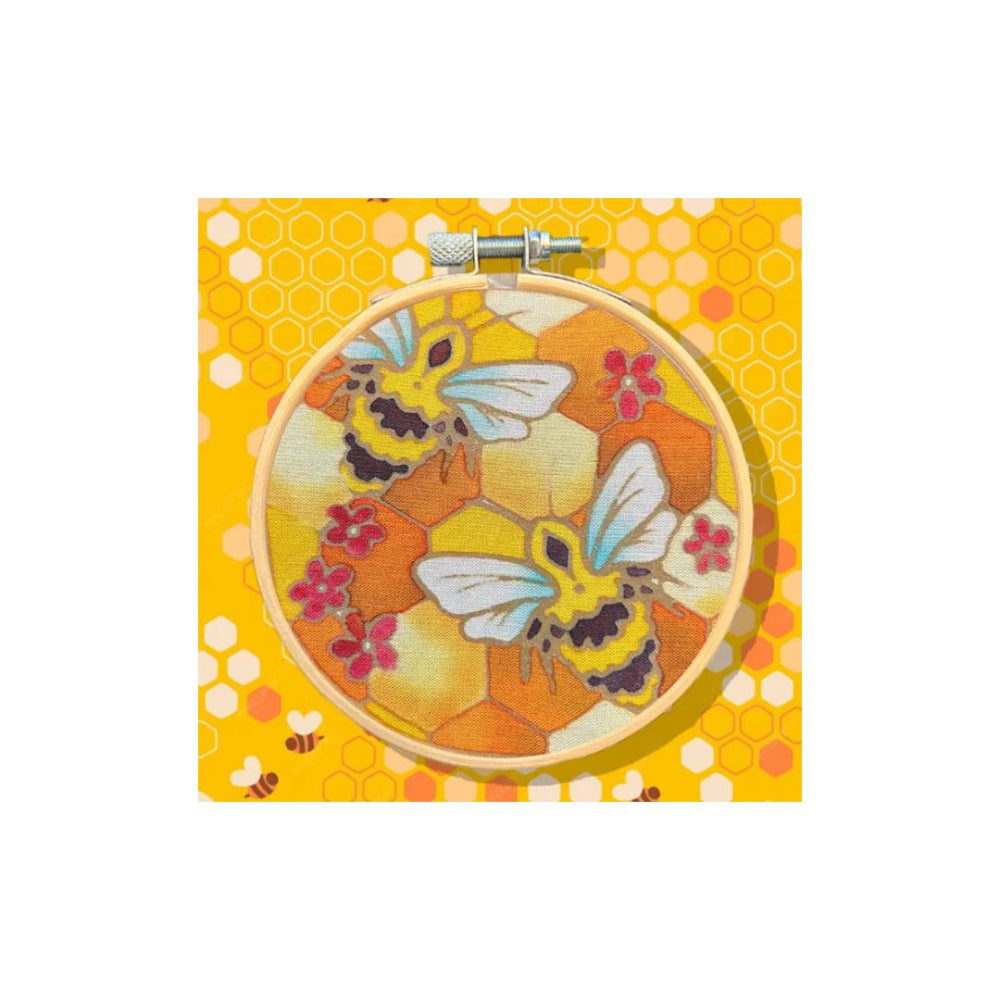 A round embroidery hoop containing a floral bee design on a yellow honeycomb-patterned fabric, shown against a matching yellow patterned background.