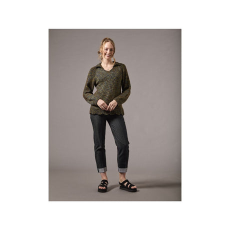 A woman modeling a dark olive green and black patterned knit sweater, paired with dark gray cuffed jeans and black sandals, standing against a solid gray studio background.