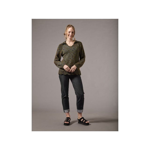 A woman modeling a dark olive green and black patterned knit sweater, paired with dark gray cuffed jeans and black sandals, standing against a solid gray studio background.