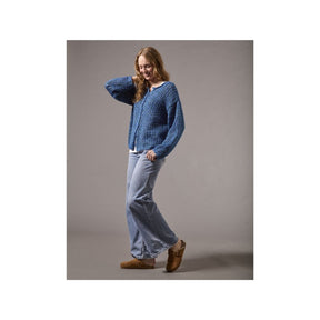 A woman modeling a blue textured knit cardigan sweater, light-wash wide-leg jeans, and brown suede mules against a solid gray studio background.