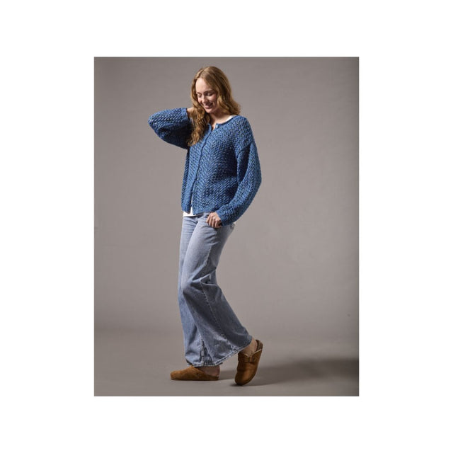 A woman modeling a blue textured knit cardigan sweater, light-wash wide-leg jeans, and brown suede mules against a solid gray studio background.