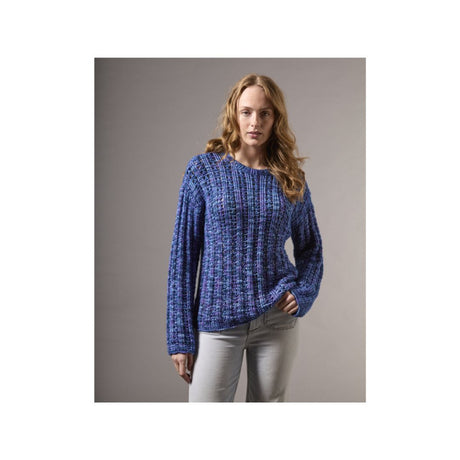 A woman modeling a textured, chunky knit sweater in a blue and purple heathered pattern, paired with light gray pants, against a solid gray studio background.