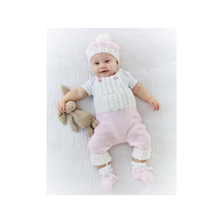 A baby lying on a white textured blanket, wearing a knitted romper, matching hat, and booties, and holding a small stuffed bunny.