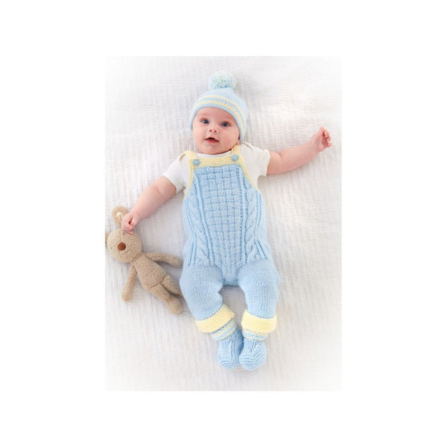 A baby lying on a white textured blanket wearing a light blue cable-knit footed romper, matching knit hat, and yellow trim, while holding a small beige stuffed bunny.