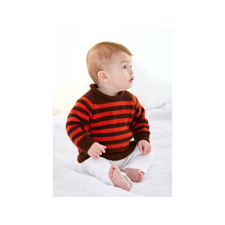A baby sitting on a white bed wearing a brown and orange striped knit sweater and white pants.