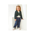 A young girl with blonde pigtails stands in a white studio setting wearing a dark green and blue textured knit cardigan over a white top, paired with dark blue jeans and white sneakers.