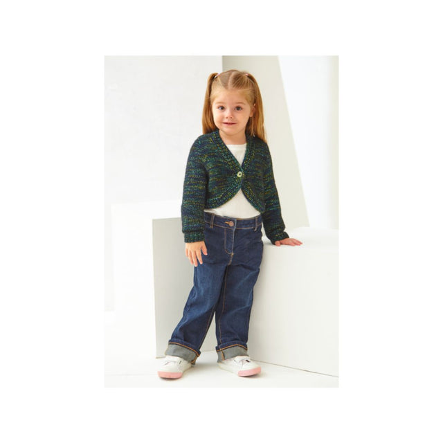 A young girl with blonde pigtails stands in a white studio setting wearing a dark green and blue textured knit cardigan over a white top, paired with dark blue jeans and white sneakers.