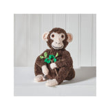A handmade brown monkey plush toy holding a small green leafy sprig, sitting on a white surface against a white paneled background.
