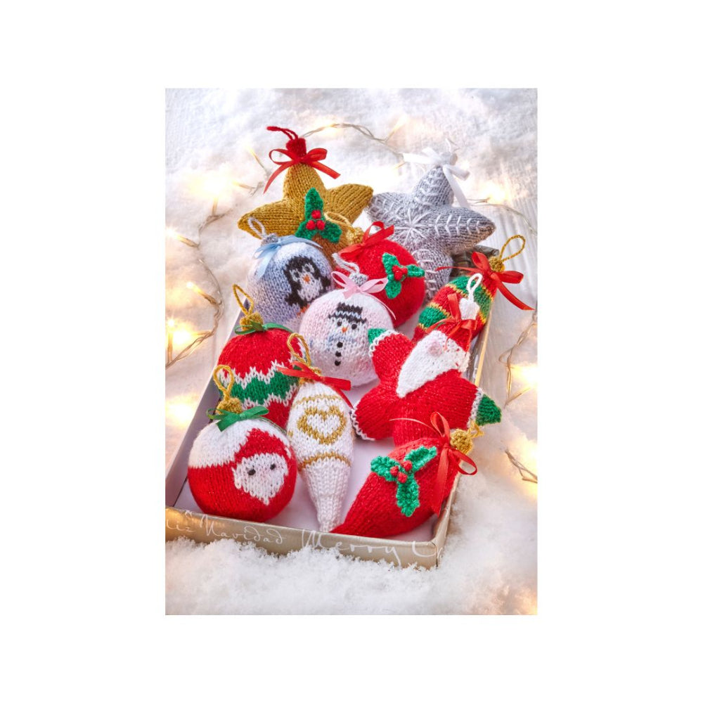 A collection of handmade knitted Christmas ornaments displayed in a gift box on a white faux-snow background with warm fairy lights. The ornaments include a gold star, a silver star, a snowman ball, a Santa Claus ball, a snow cone, and a red stocking.