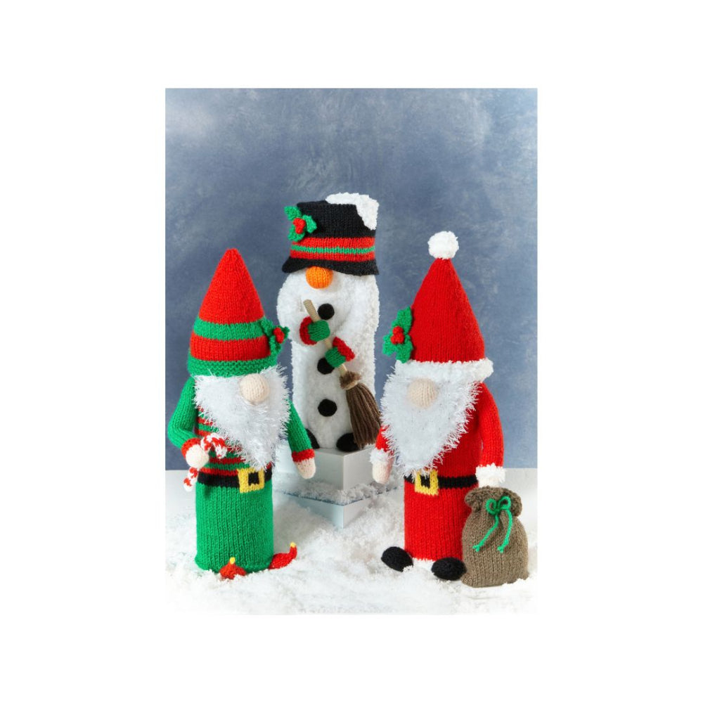 Three knitted Christmas holiday figures standing in a snowy display: an elf on the left in red and green, a snowman in the center with black buttons and a brown broom, and a Santa-style gnome on the right in red with a brown sack, set against a blue-gray snowy backdrop.
