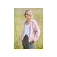 A woman standing in a grassy field wearing a light pink chunky knit cardigan over a white top and olive green trousers.