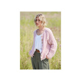 A woman standing in a grassy field wearing a light pink chunky knit cardigan over a white top and olive green trousers.