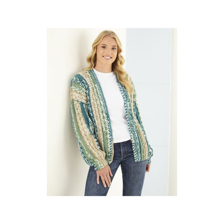A woman wearing a striped open-front cardigan with green, teal, beige, and white patterns, layered over a white t-shirt and blue jeans, standing against a light-coloured wall.