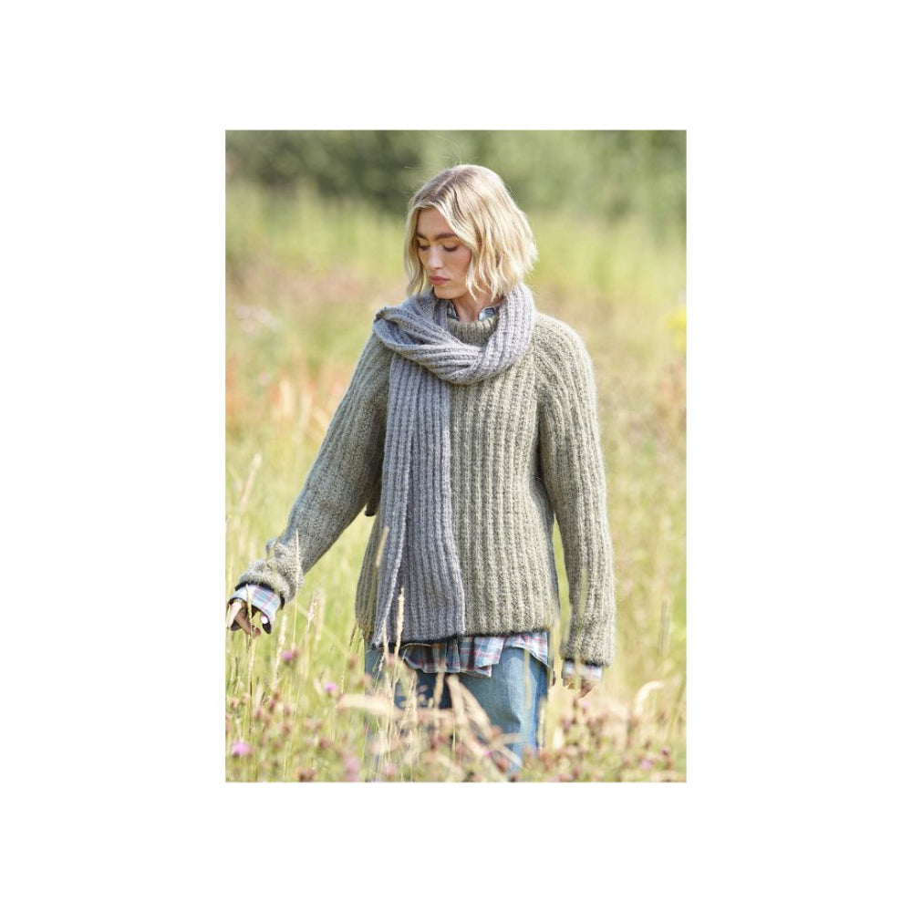 A woman standing in a grassy field wearing a chunky ribbed knit sweater and matching scarf in a muted grey-green colour, with blue jeans and a plaid shirt visible.
