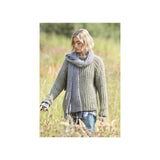 A woman standing in a grassy field wearing a chunky ribbed knit sweater and matching scarf in a muted grey-green colour, with blue jeans and a plaid shirt visible.