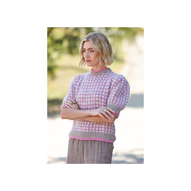 A woman wearing a pink and white patterned knit sweater with puff sleeves and a ribbed hem, paired with brown trousers, standing outdoors with her arms crossed.