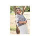 A woman stands outdoors leaning against a tree trunk, wearing a chunky knit short-sleeve sweater with horizontal grey and white stripes and white pants.