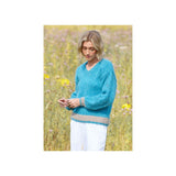A woman standing in a sunlit field of wildflowers, wearing a fuzzy turquoise blue sweater with a beige ribbed hem and cuffs, paired with white pants.