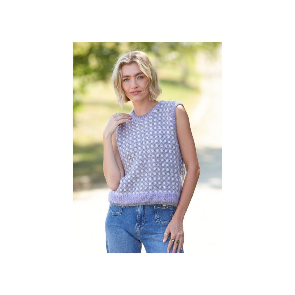 A woman wearing a lavender sleeveless knit sweater vest with a white geometric pattern, paired with blue jeans, standing outdoors against a blurred green background.