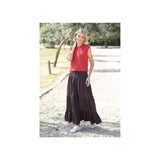A woman stands outdoors on a gravel path wearing a sleeveless red knit top and a long dark brown tiered skirt with white sneakers.