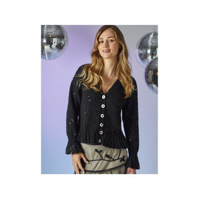 A woman modeling a black knit cardigan with white buttons, featuring a V-neck and ruffled cuffs, set against a lavender background with hanging disco ball decorations.