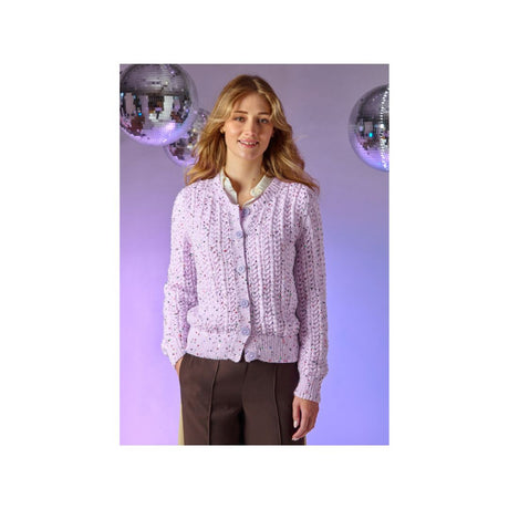 A woman wearing a light purple lace cardigan with button-front closure, paired with dark brown trousers, standing against a lavender studio backdrop with mirrored disco ball props.