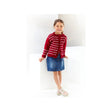 A young girl stands in a bright white studio setting, wearing a red knit cardigan with a white Nordic-style pattern, a blue denim skirt, and light pink sneakers.