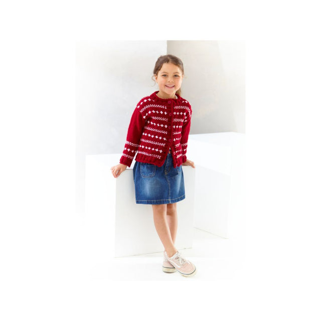 A young girl stands in a bright white studio setting, wearing a red knit cardigan with a white Nordic-style pattern, a blue denim skirt, and light pink sneakers.