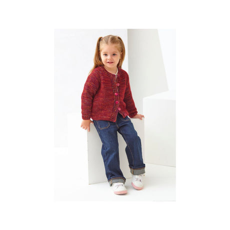 A young girl with blonde pigtails sits on a white geometric block, wearing a red and blue textured knit cardigan, blue jeans, and white sneakers.
