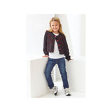A young girl with blonde hair stands in a white geometric studio setting, wearing a dark patterned cardigan with pink buttons over a white t-shirt, blue jeans, and white sneakers.