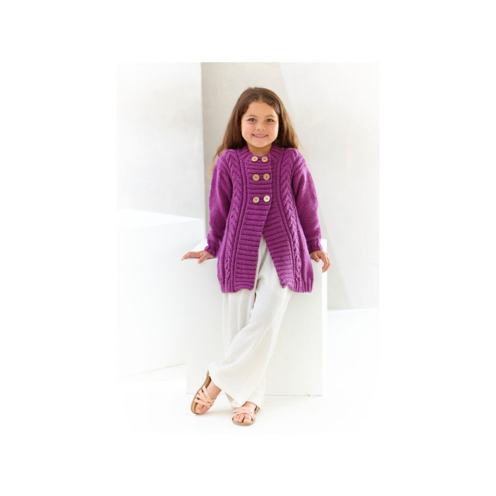 A young girl wearing a purple cable-knit cardigan with gold buttons, white pants, and sandals, standing in a bright white studio setting.